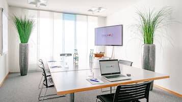 A conference room with a long table, chairs, a laptop, a TV screen, and two large potted plants.