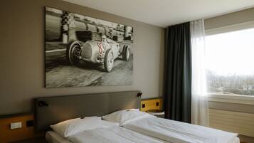 A hotel room with a bed, a headboard, a nightstand, a wall-mounted painting of a vintage race car, and a window with curtains.