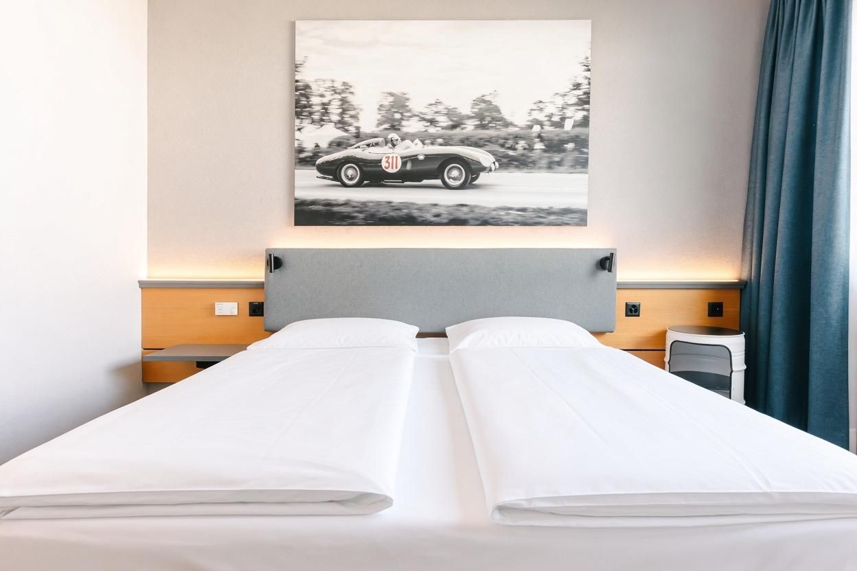 A hotel room with a bed, a headboard, a nightstand, a chair, and a wall-mounted picture of a vintage car.