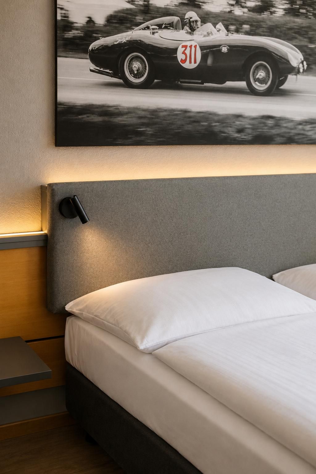 Une chambre à coucher comprenant un lit, une tête de lit, une table de chevet et un tableau représentant une voiture ancienne, accroché au mur.