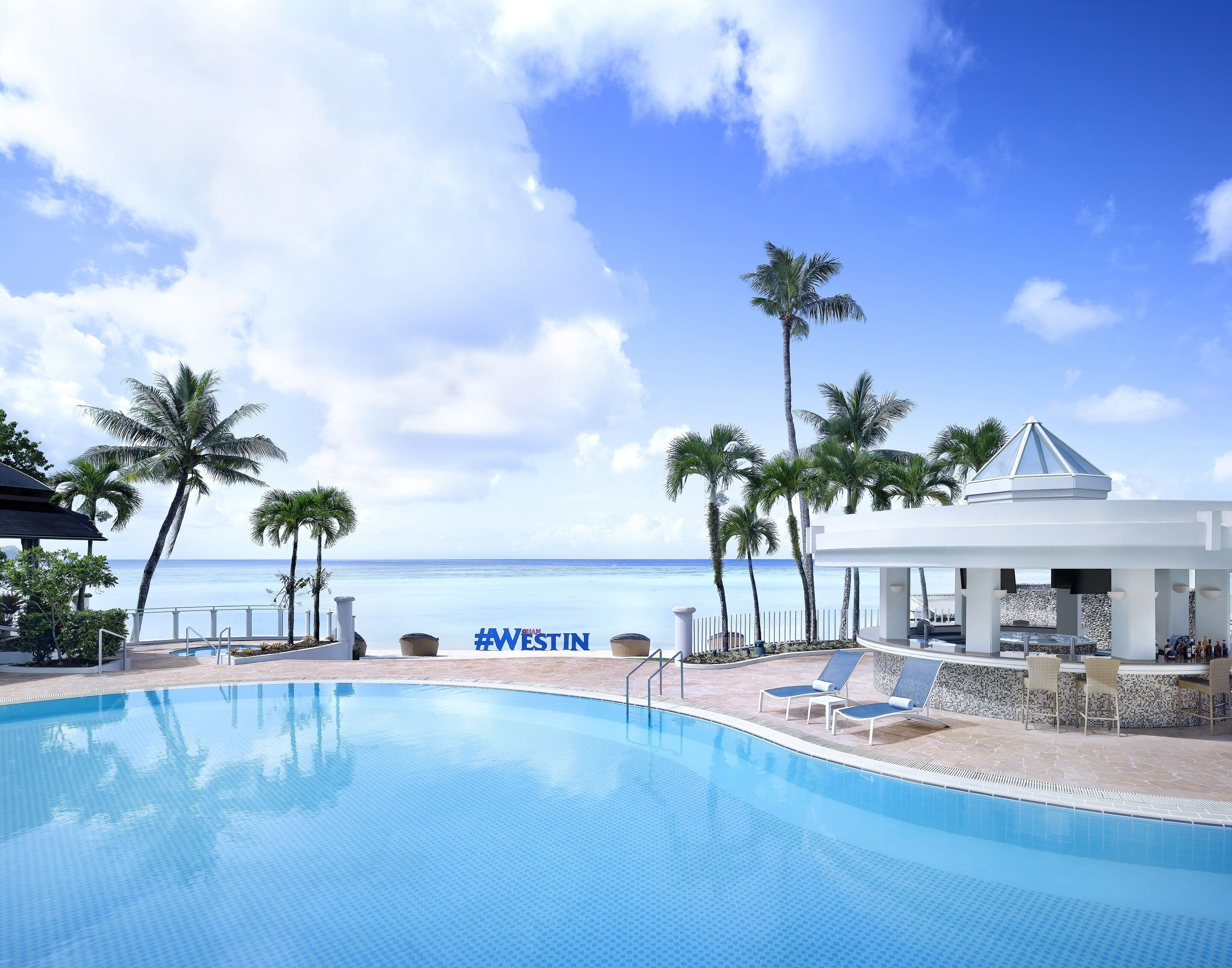 A pool area with a view of the ocean, palm trees, and a covered seating area.