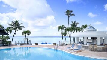 A pool area with a view of the ocean, palm trees, and a covered seating area.