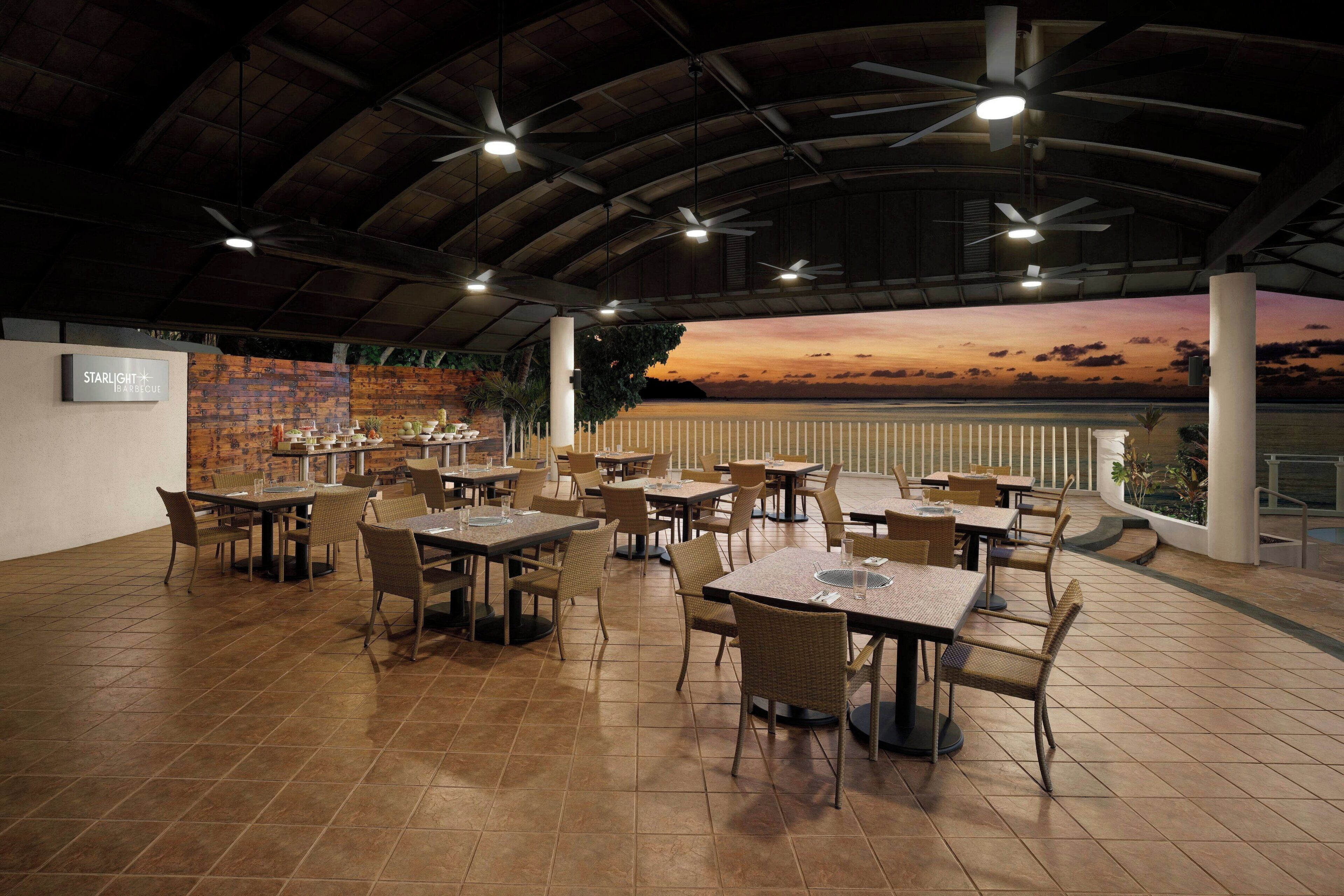 A spacious indoor dining area with multiple tables and chairs, a high ceiling with fans, and a view of the sunset over the water.