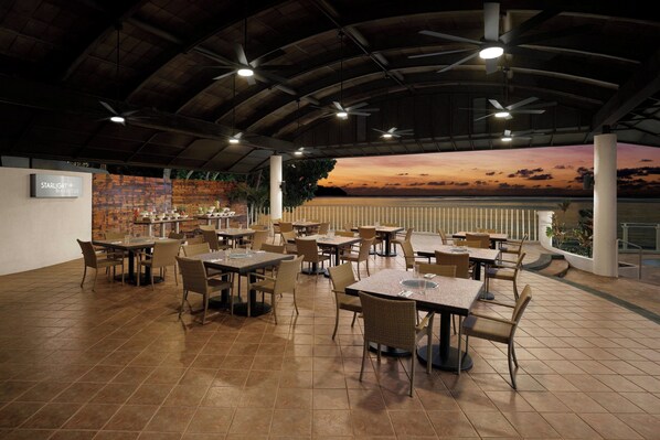 A spacious indoor dining area with multiple tables and chairs, a high ceiling with fans, and a view of the sunset over the water.