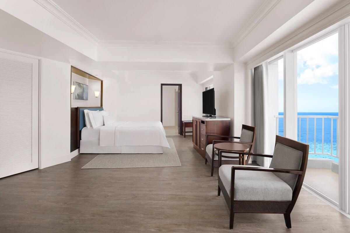 A hotel room with a bed, a desk, a chair, and a large window with a view of the ocean.