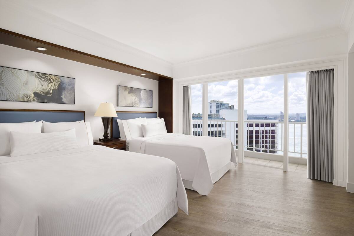 A hotel room with two beds, a nightstand, a lamp, and a large window with a city view.