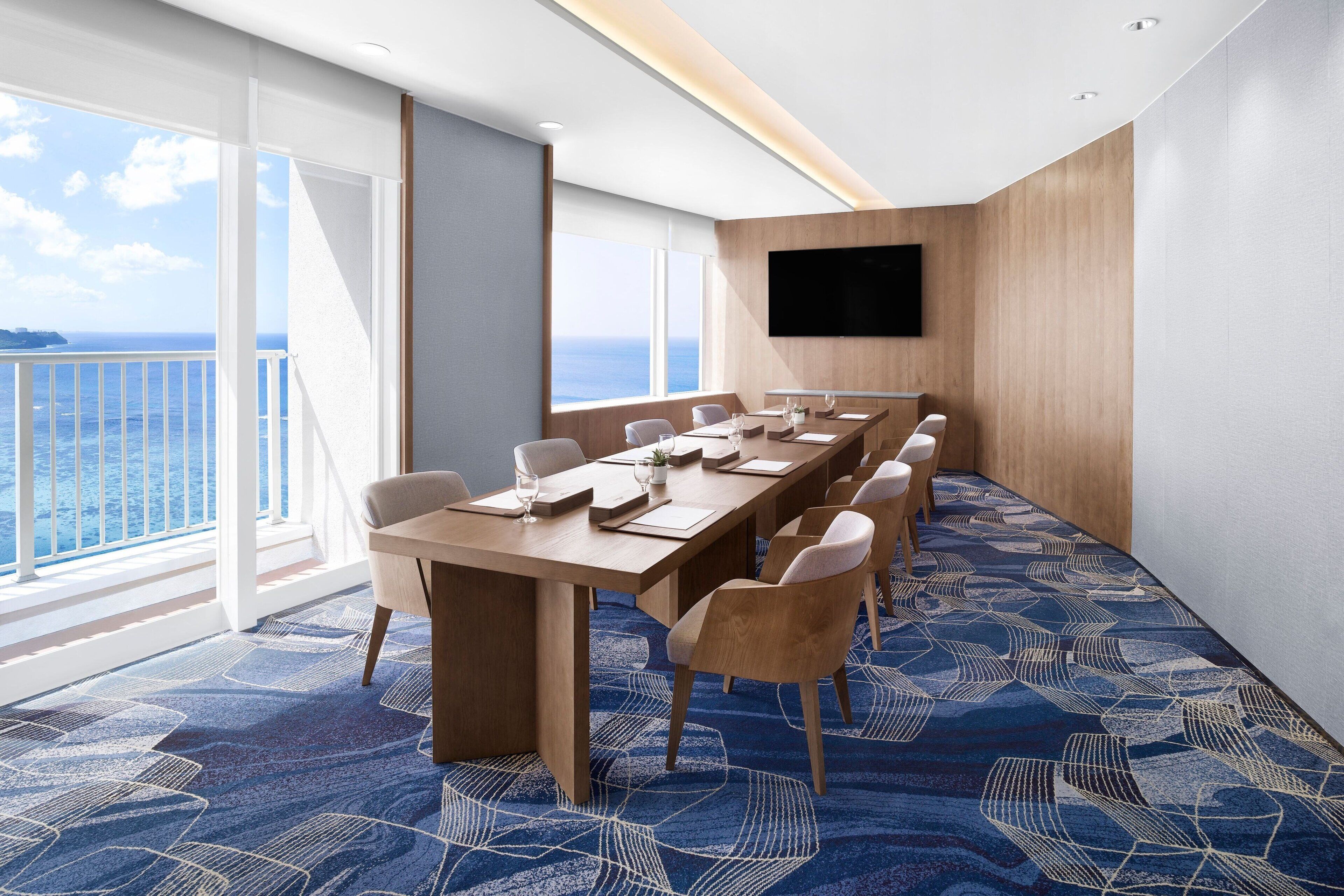 A conference room with a long table, chairs, a flat-screen TV, and a large window with a view of the sea.