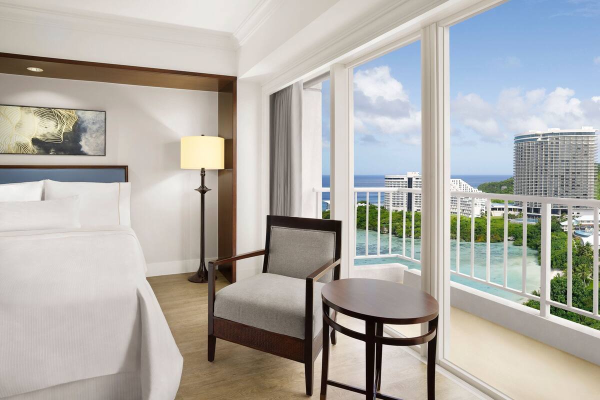A hotel room with a bed, a chair, a table, a lamp, and a large window offering a view of a cityscape and ocean.