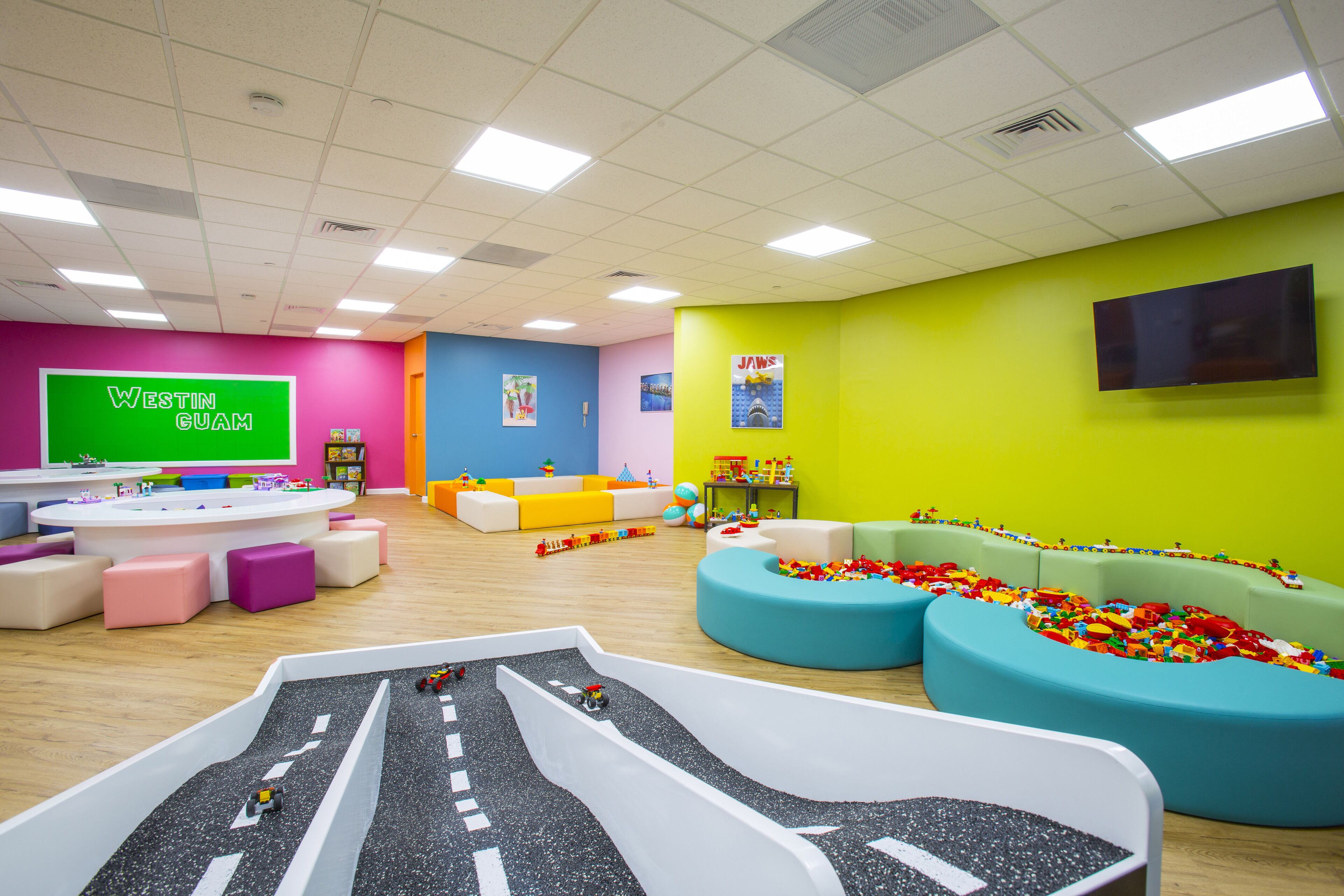 A brightly colored playroom with a slide, colorful seating areas, and a green screen displaying "WESTIN GUAM".