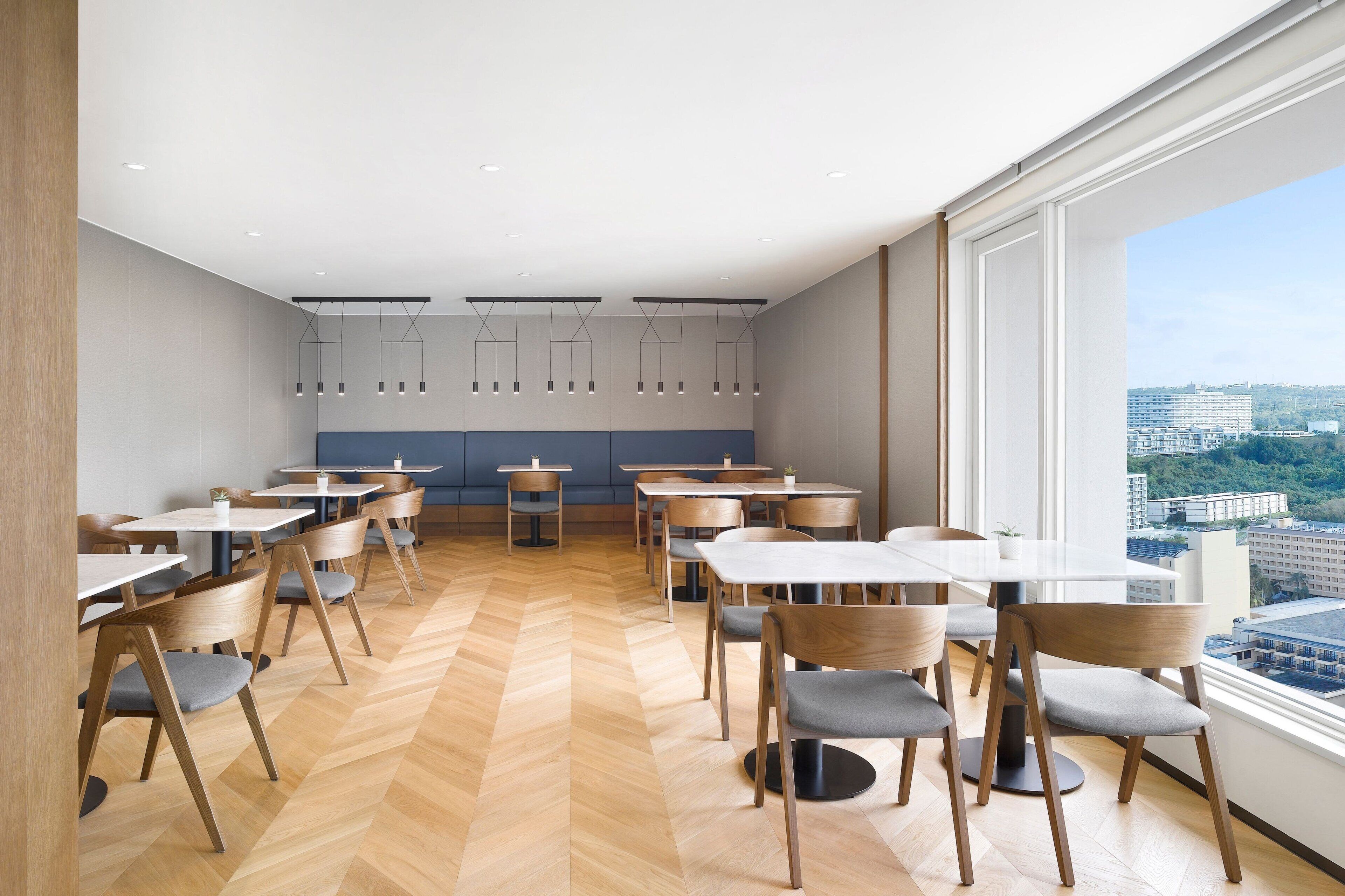 A modern dining area with wooden chairs and tables, a herringbone floor, and large windows offering a city view.