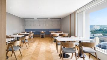 A modern dining area with wooden chairs and tables, a herringbone floor, and large windows offering a city view.