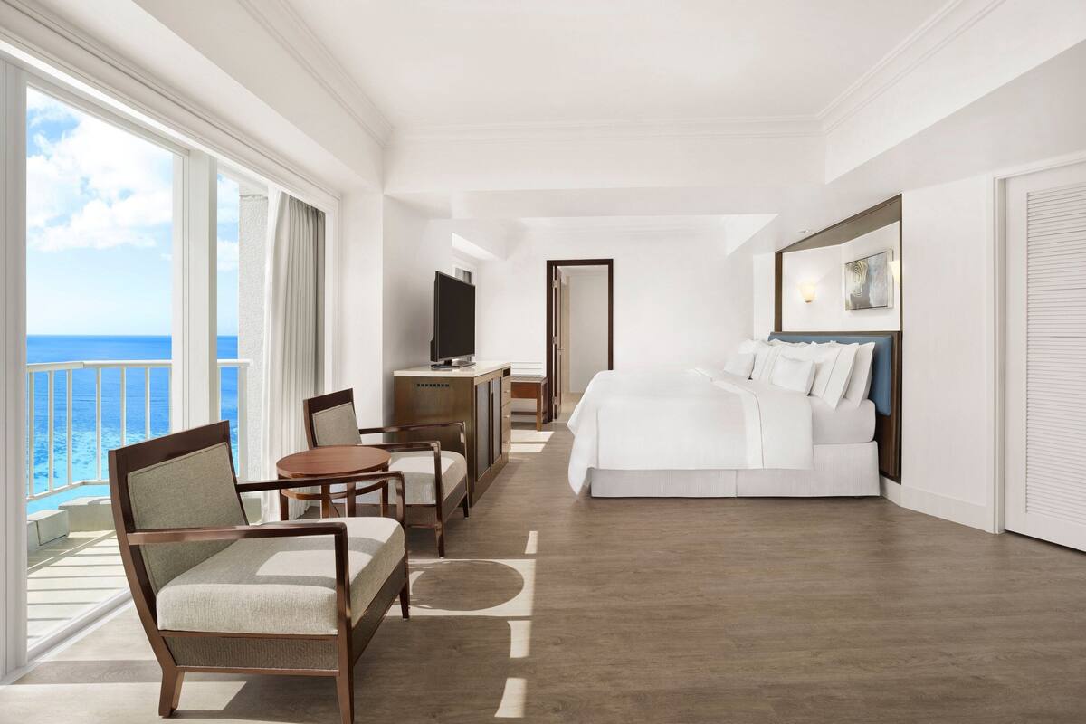 A hotel room with a large bed, a desk, a chair, a television, and a balcony with a view of the ocean.