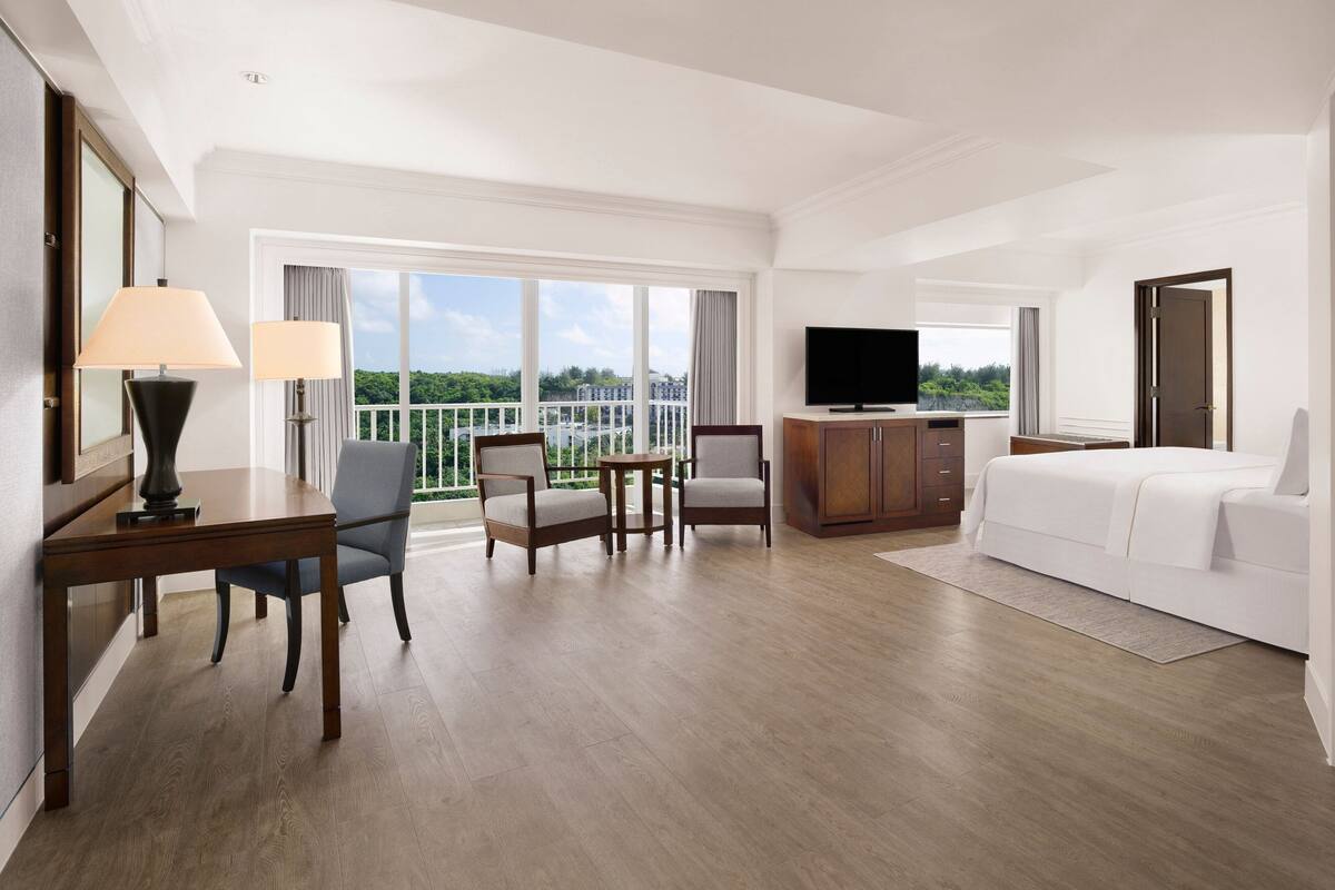 A hotel room with a large bed, a TV, a desk with a lamp, two chairs, a small table, and a balcony with a view.