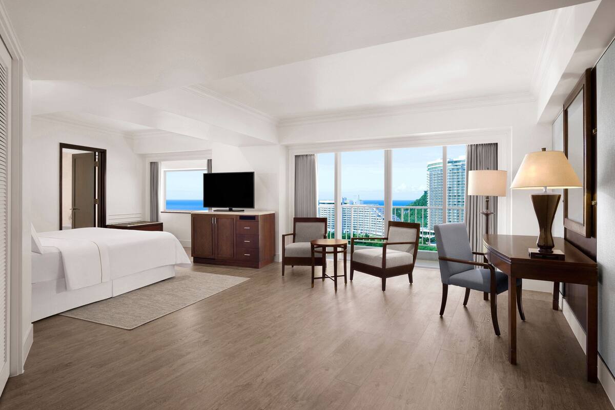 A hotel room with a bed, a desk, a chair, a television, and large windows offering a city view.