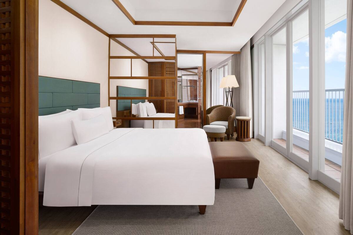 A hotel room with a large bed, a seating area, a small table, and a view of the sea.
