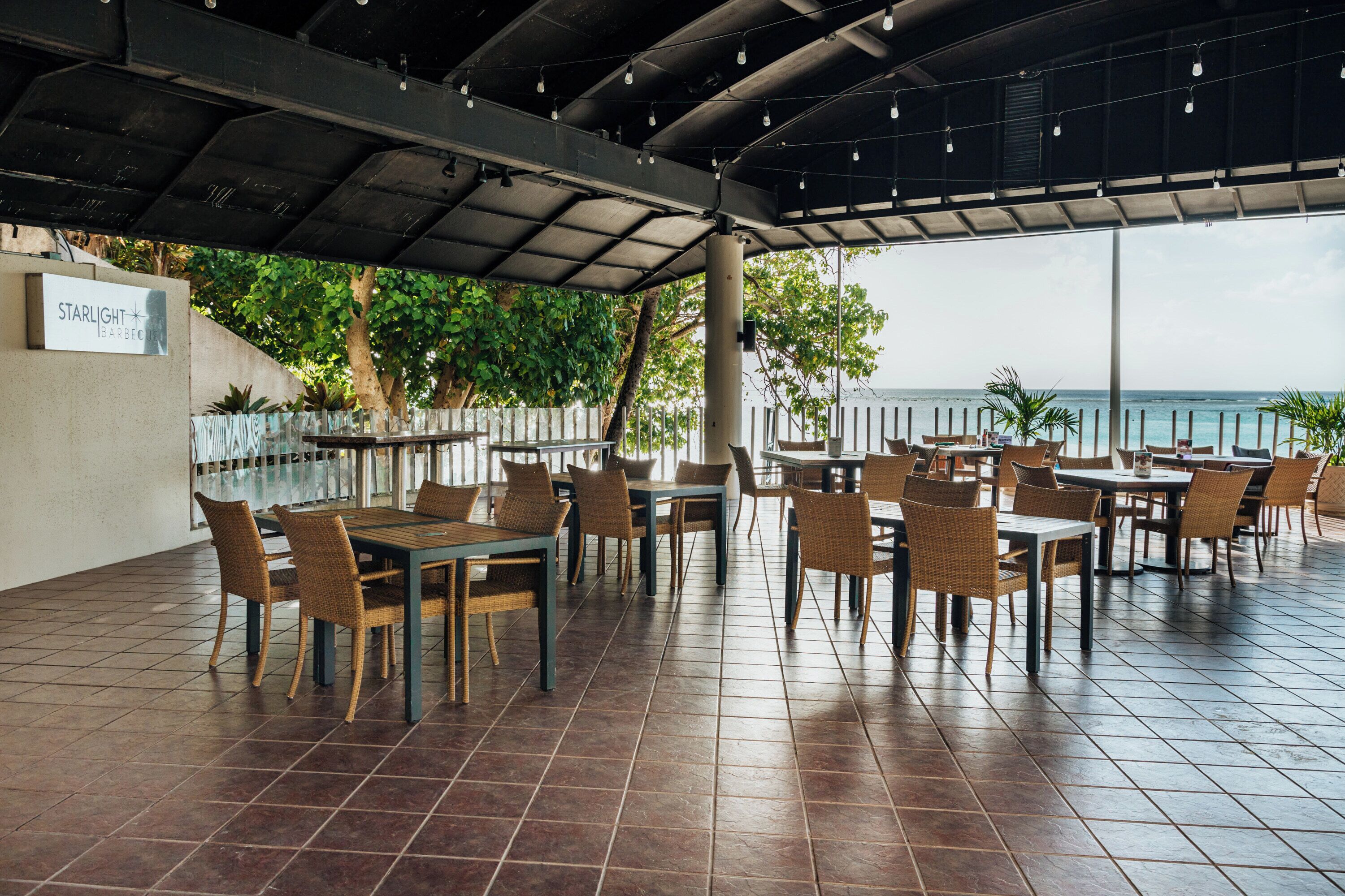 An outdoor restaurant with multiple tables and chairs, a covered area with hanging lights, and a view of the ocean.