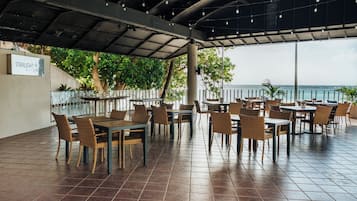 An outdoor restaurant with multiple tables and chairs, a covered area with hanging lights, and a view of the ocean.