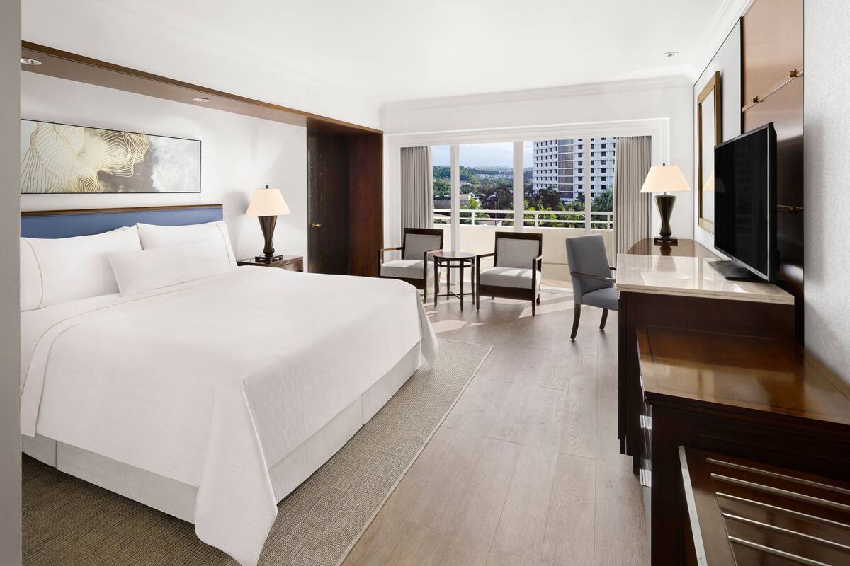 A hotel room with a large bed, two armchairs, a small table, a TV, and a view of the city through large windows.
