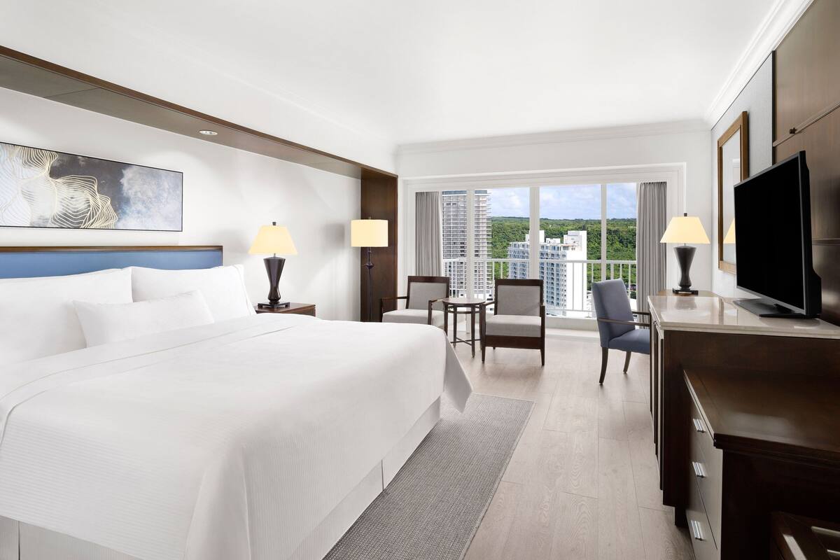 A hotel room with a large bed, two bedside tables with lamps, a TV, a desk with a chair, and a balcony with a view of buildings.