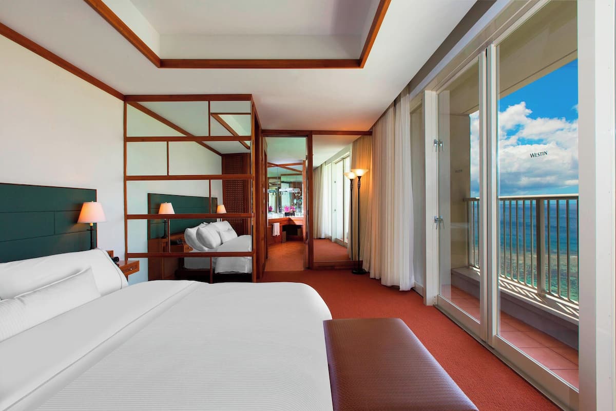 A hotel room with a large bed, a desk, a chair, a balcony with a view of the sea, and a sliding door leading to a bathroom.