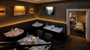 A dimly lit dining area with round tables, dark upholstered booths, and wall-mounted lighting.
