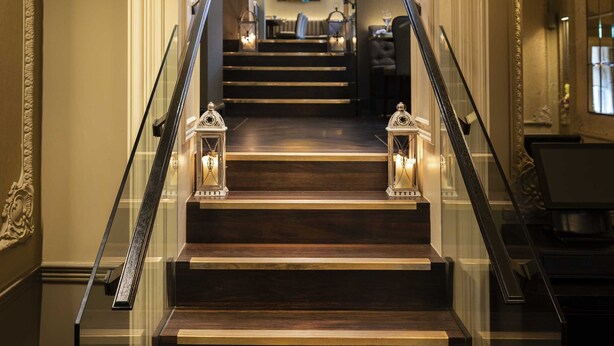 A grand staircase with glass railings and ornate wall decorations, leading to a well-lit room with a chandelier.