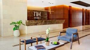 Un espace d’accueil d’hôtel comprenant un comptoir de réception, un coin salon avec des chaises, une plante en pot et un mur décoratif portant le nom de l’hôtel.
