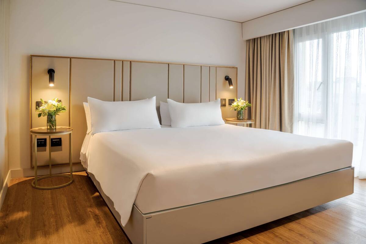 Una habitación de hotel moderna con una cama grande, dos mesitas de noche con lámparas y vistas al exterior a través de cortinas translúcidas.