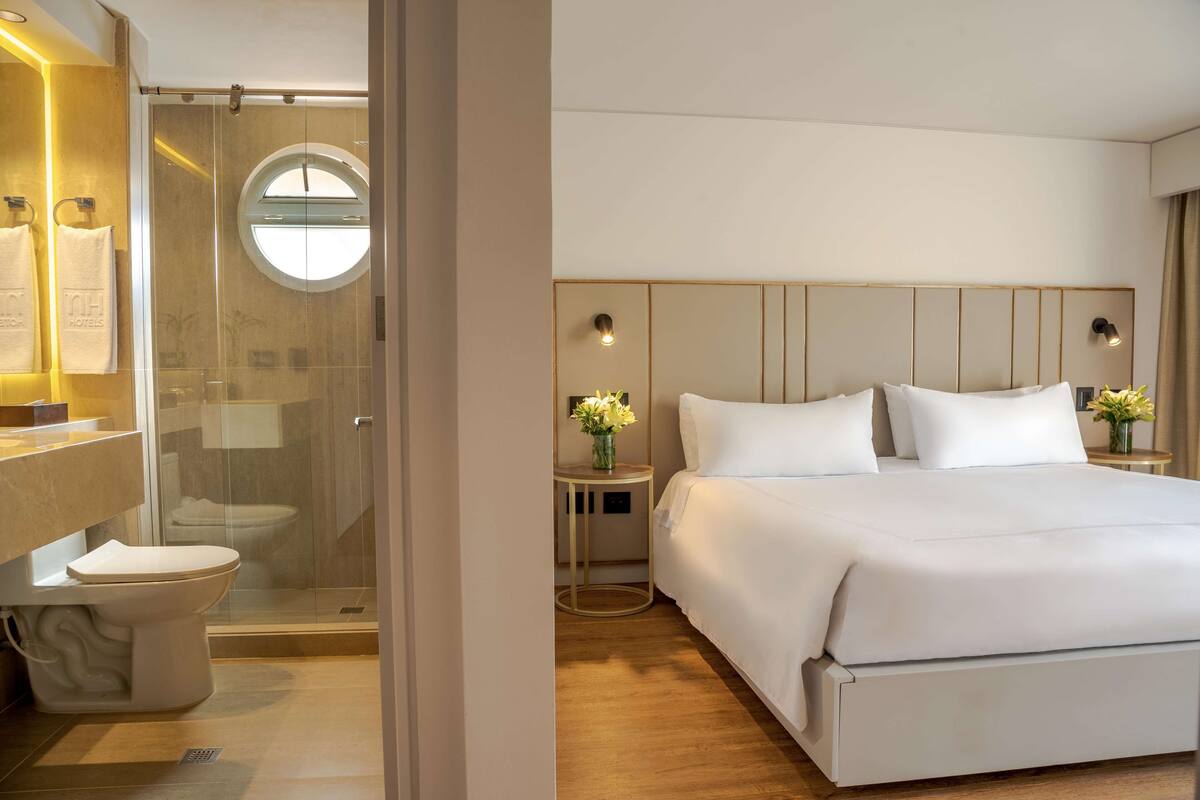 Habitación de hotel con una cama grande, un ventanal redondo, cabina de ducha con paredes de vidrio y una mesita de noche con un florero.