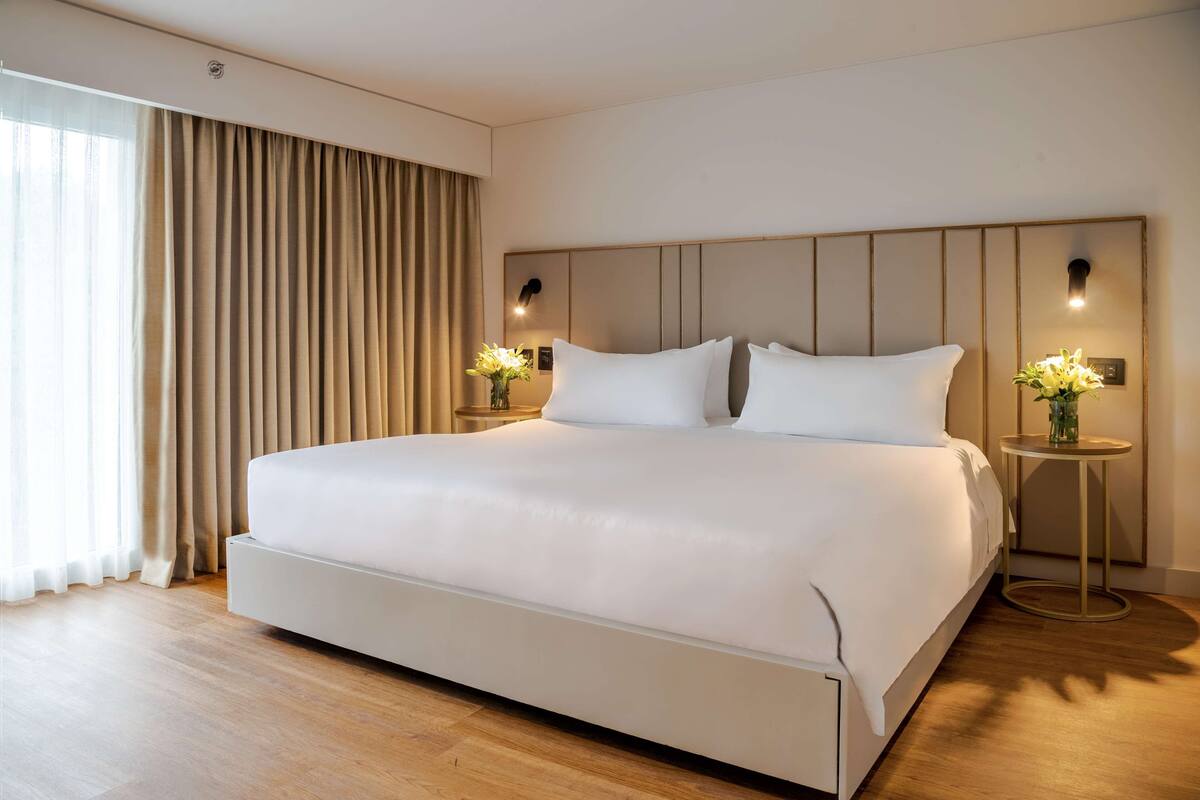 Habitación de hotel moderna con una cama grande, suelo de madera y vistas al exterior a través de cortinas traslúcidas.