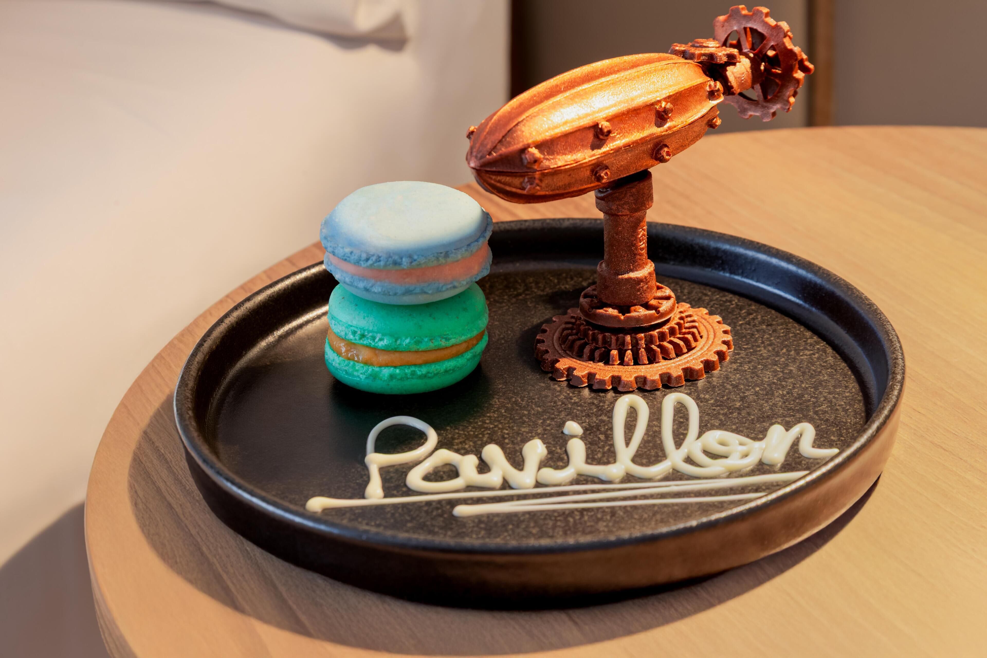 A tray with macarons and a chocolate gear, and the word 'Pavillon' written on it.