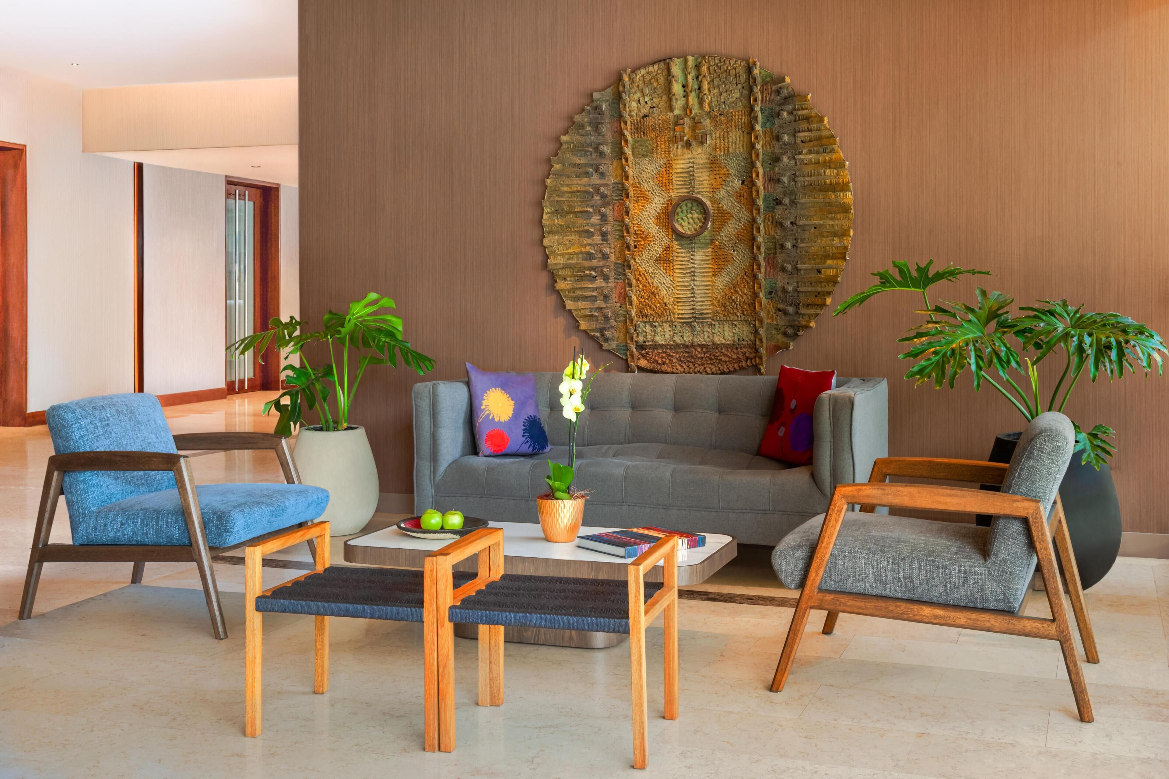 Un salon moderne avec un canapé gris, des fauteuils bleus, une table basse et deux tables d’appoint. On y trouve également des plantes en pot et un élément décoratif mural.