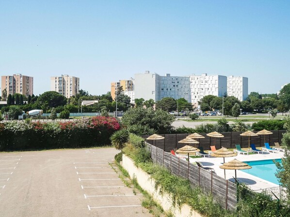 Un estacionamiento, una piscina con sillones de descanso y edificios residenciales al fondo.