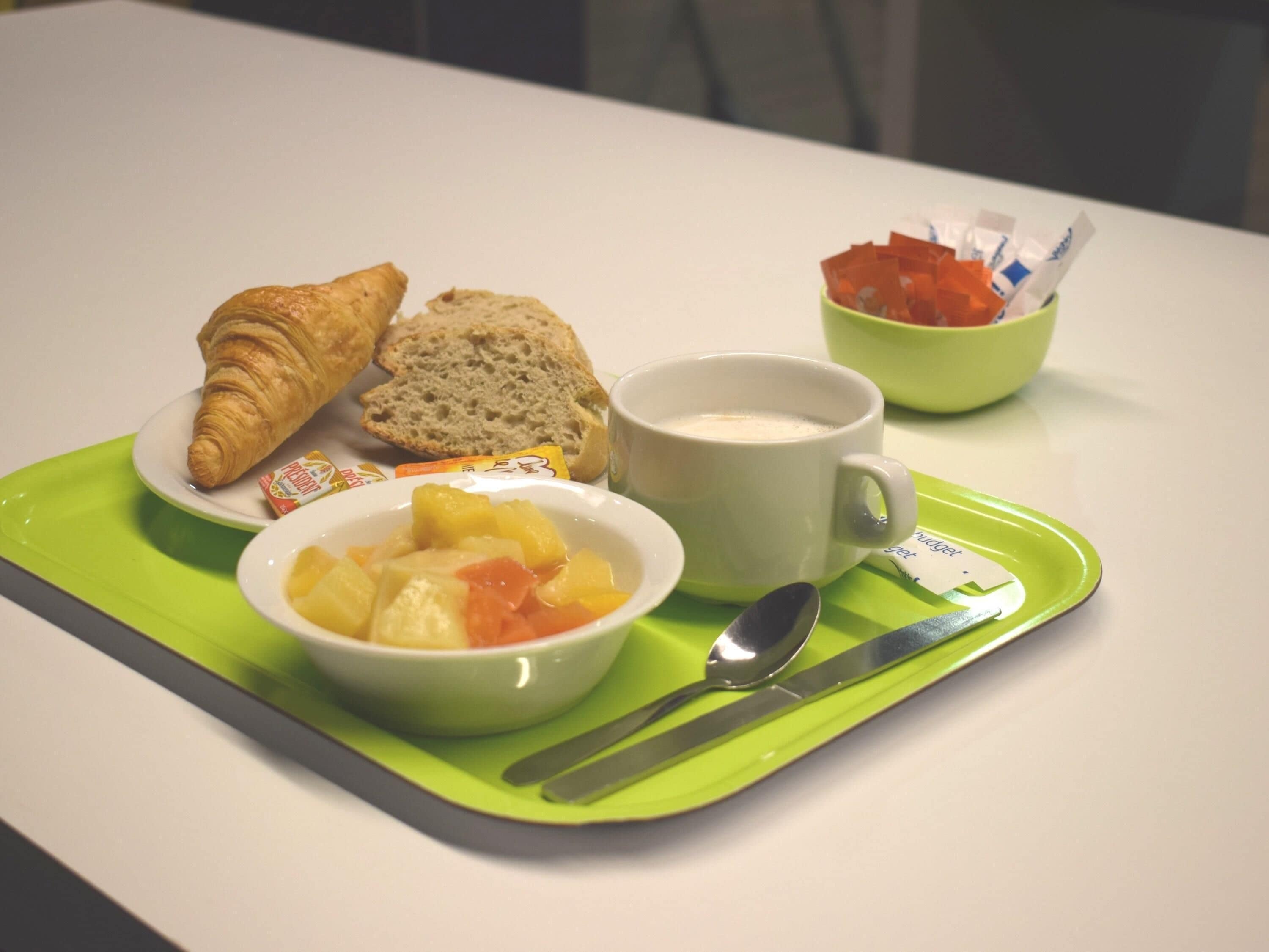 A breakfast tray with croissant, bread, fruit salad, a mug, and condiments.