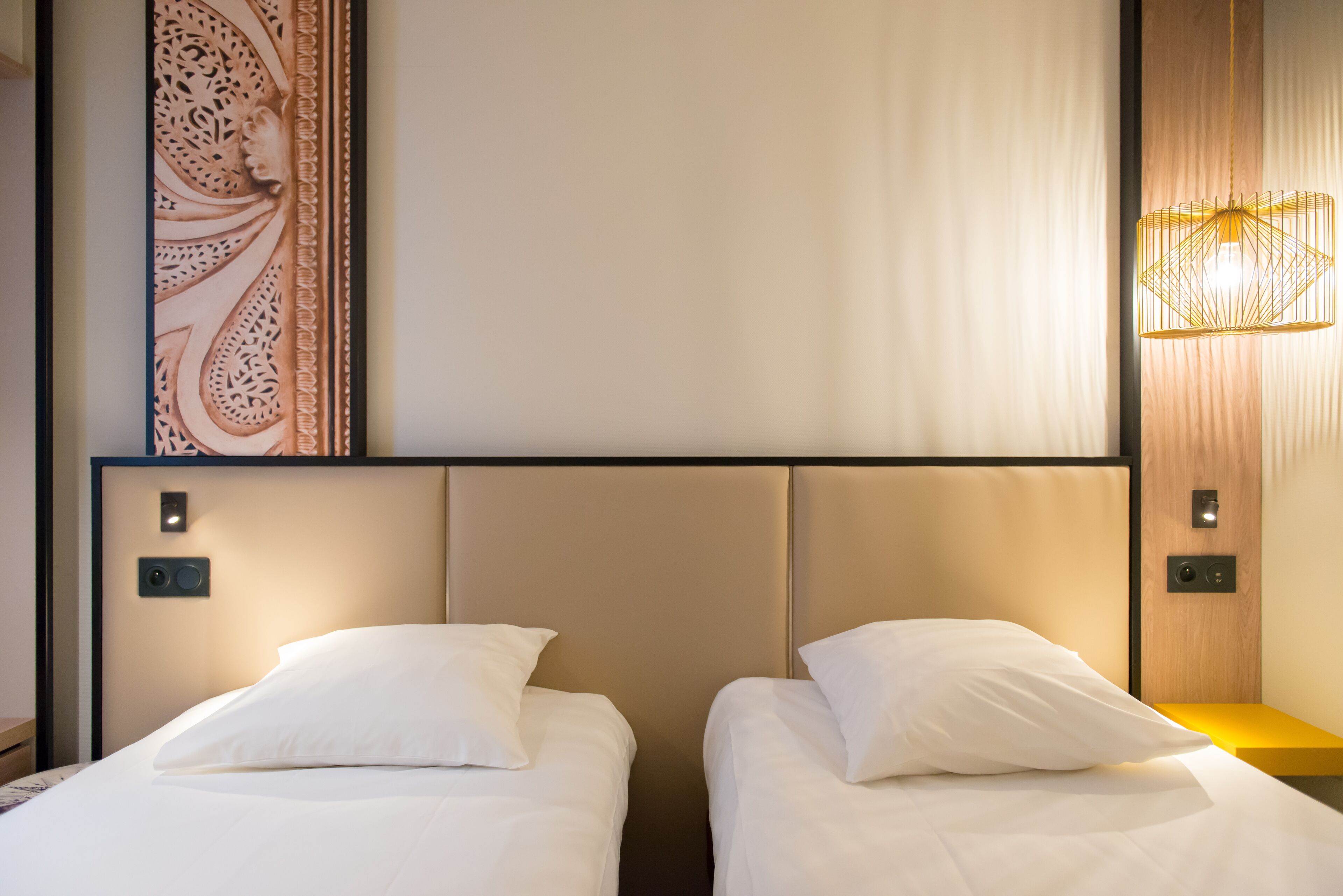Quarto de hotel com duas camas, um painel decorativo e uma luminária moderna.