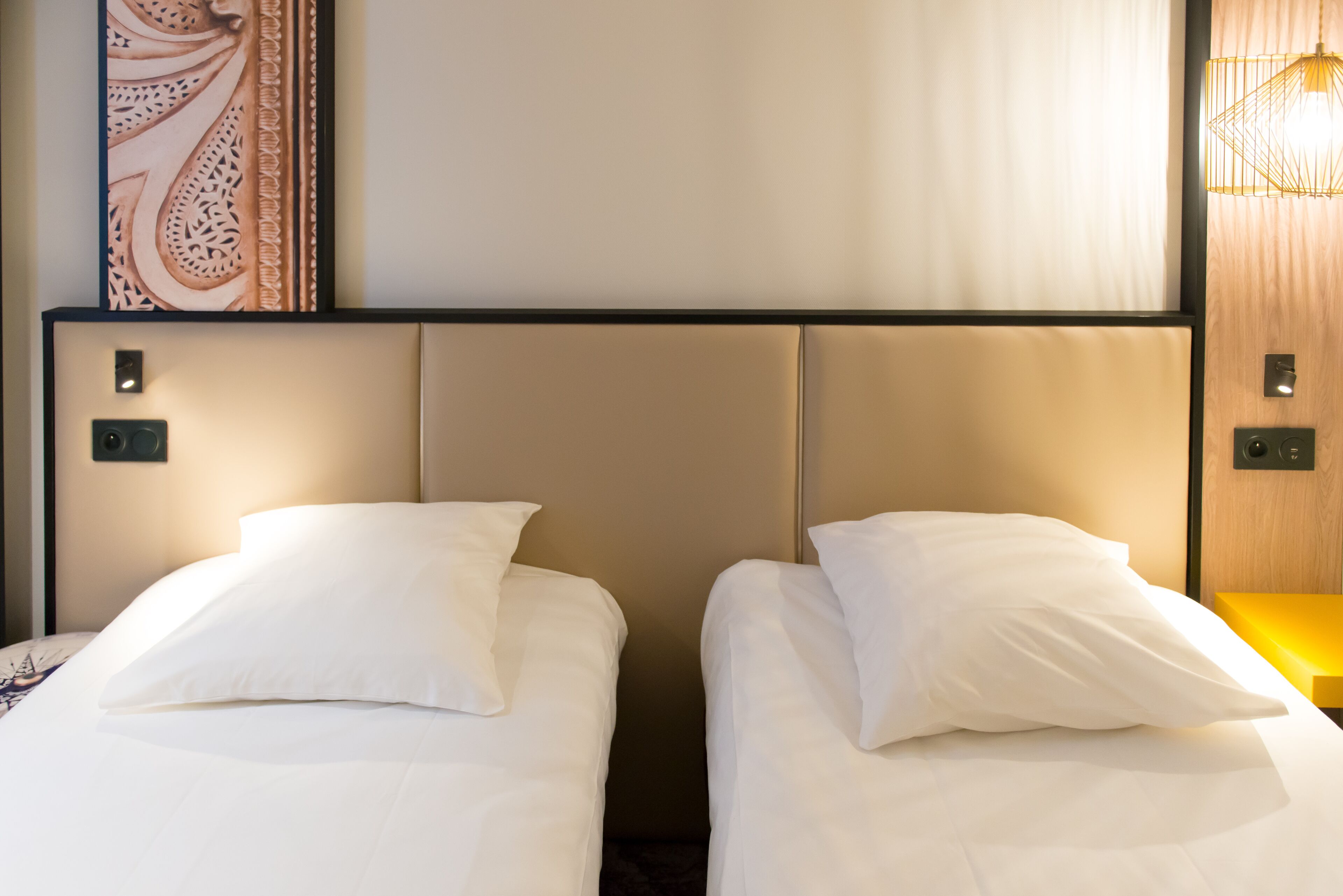 Two single beds with white linens and headboards, a wall-mounted artwork, and a modern light fixture.