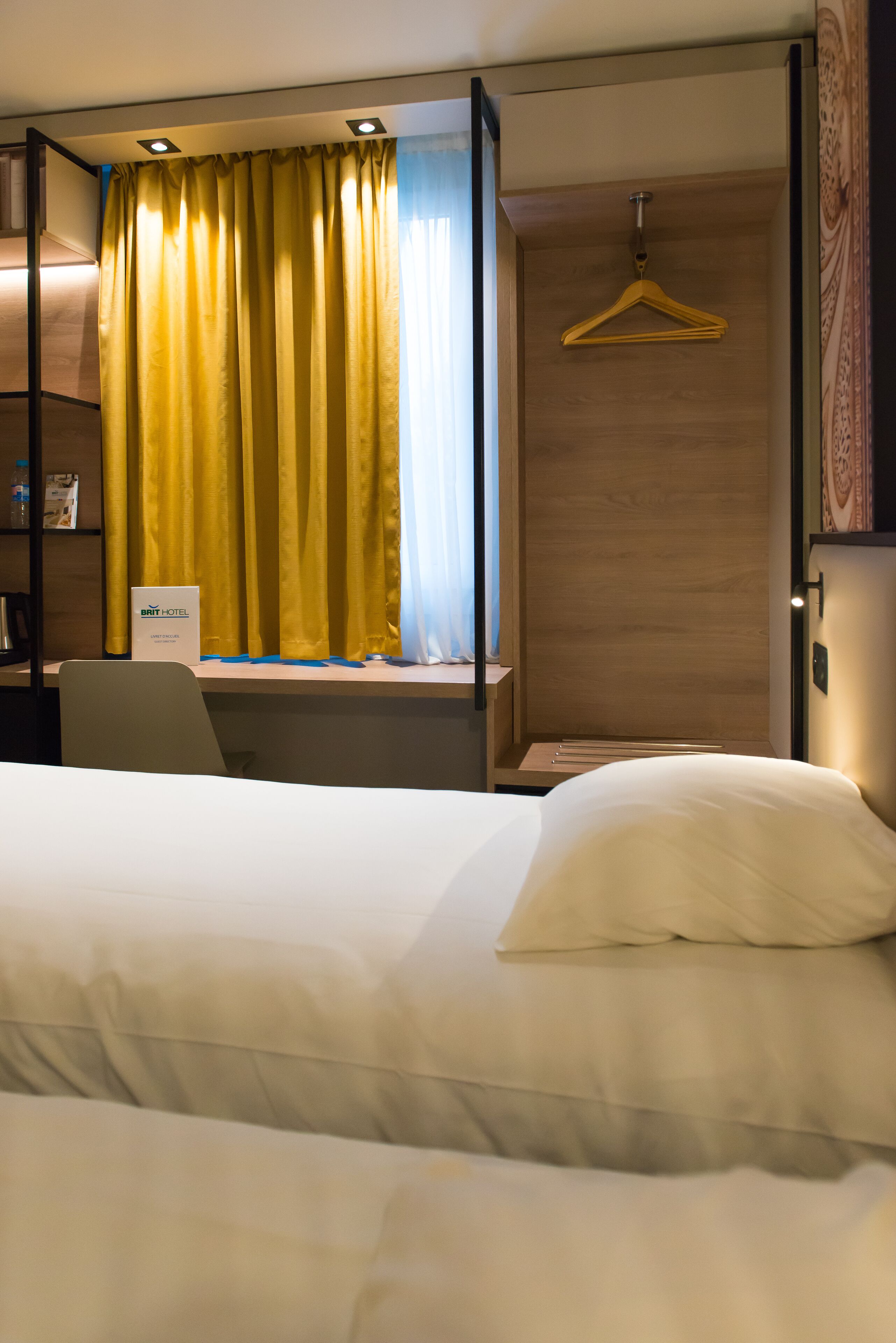 Quarto de hotel com cama, escrivaninha, cadeira, cabideiro e placa com o nome do hotel.