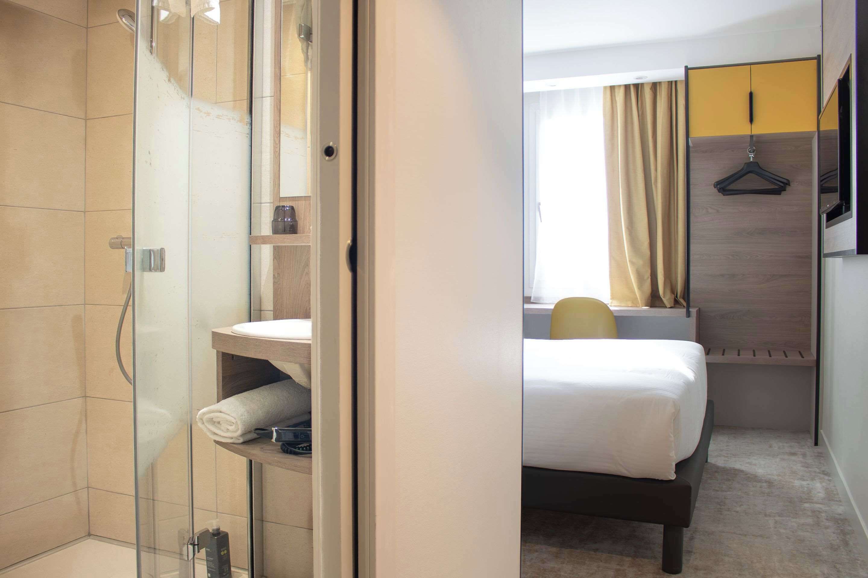 Quarto de hotel com box de vidro, cama com colchão branco, mesa de cabeceira e um cabideiro.