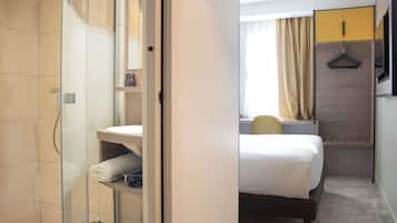 Quarto de hotel com box de vidro, cama com colchão branco, mesa de cabeceira e um cabideiro.