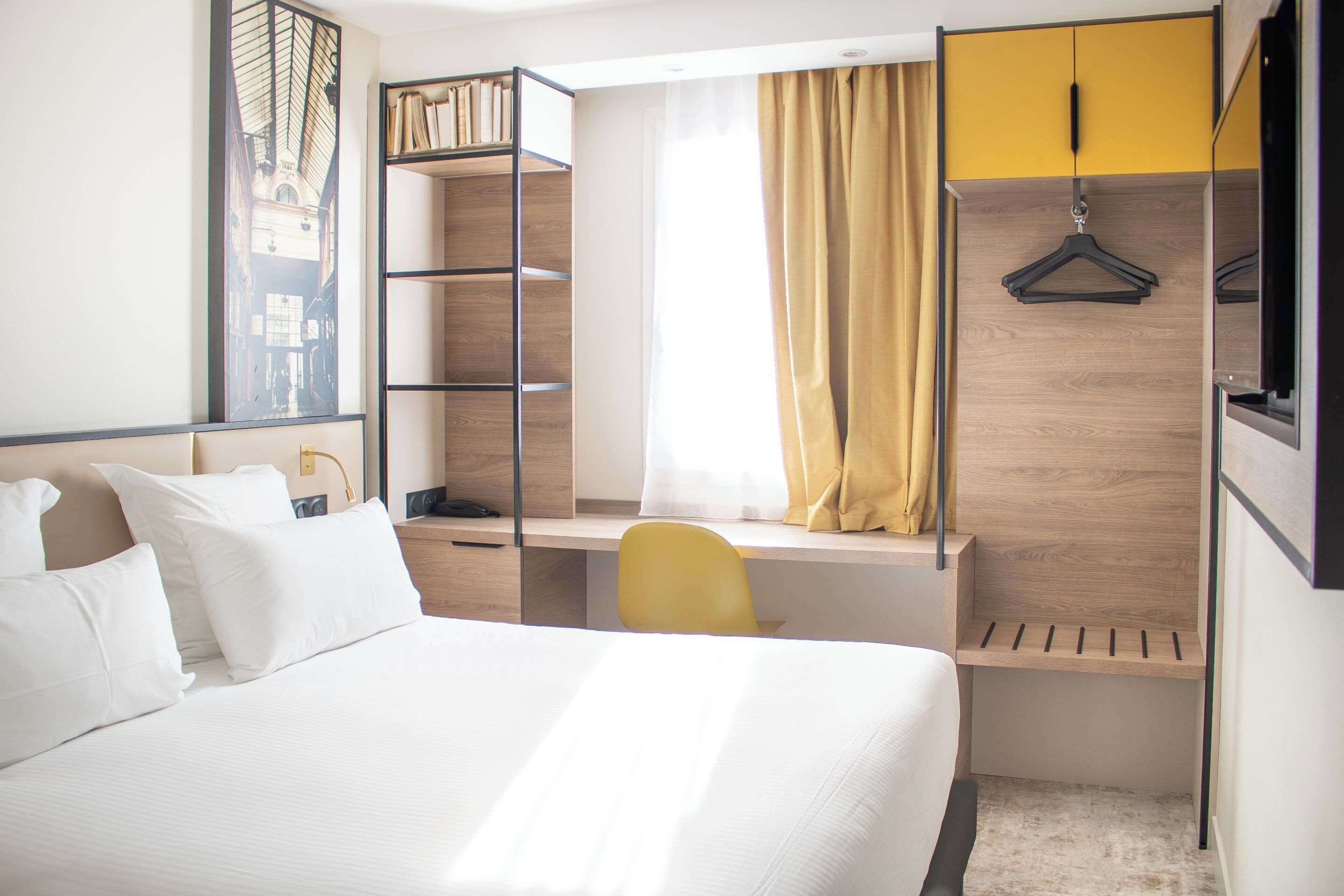 A hotel room with a bed, a desk with a chair, a wardrobe, and a window with curtains.