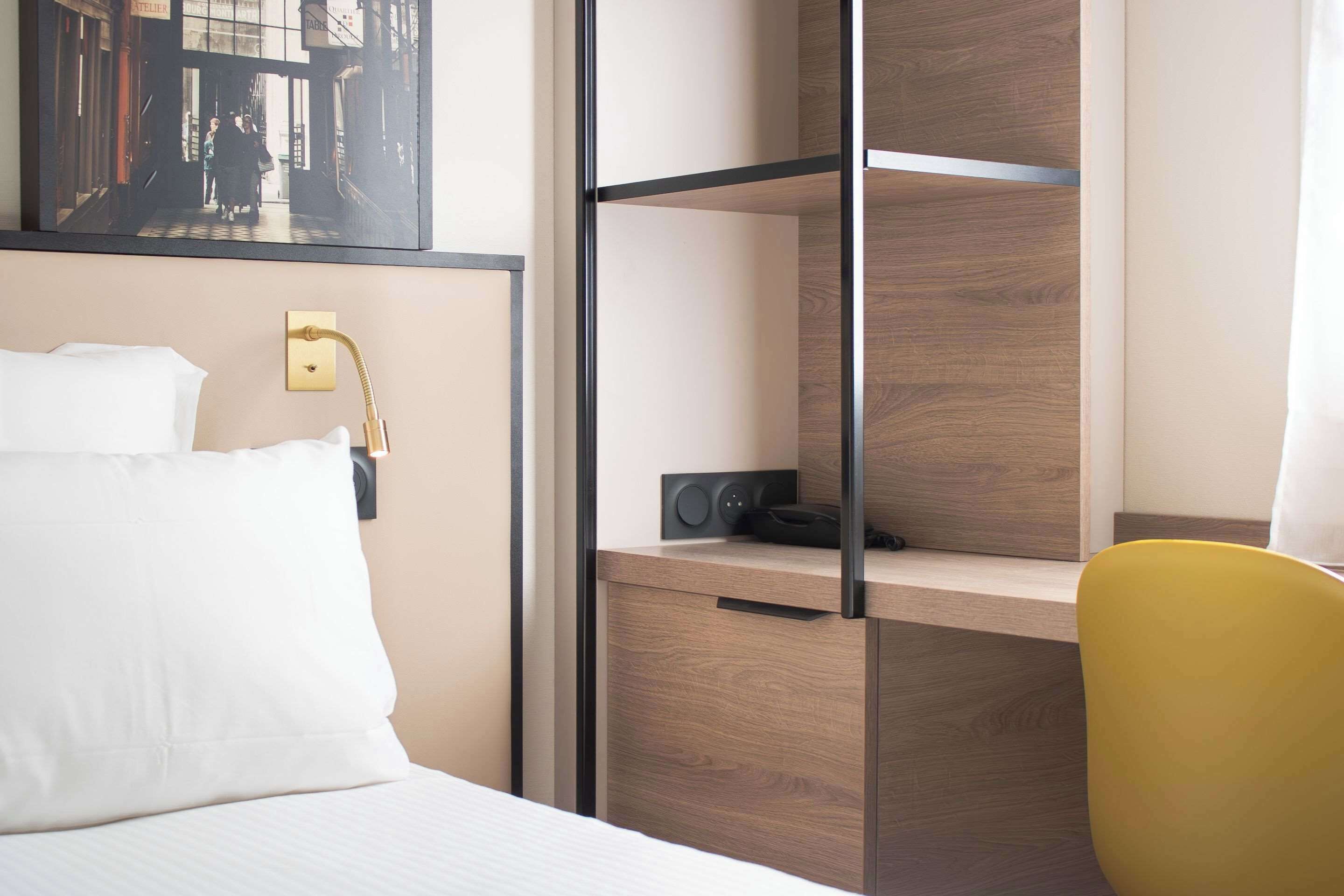 A hotel room with a bed, a desk with a chair, a wall-mounted lamp, and a framed picture.