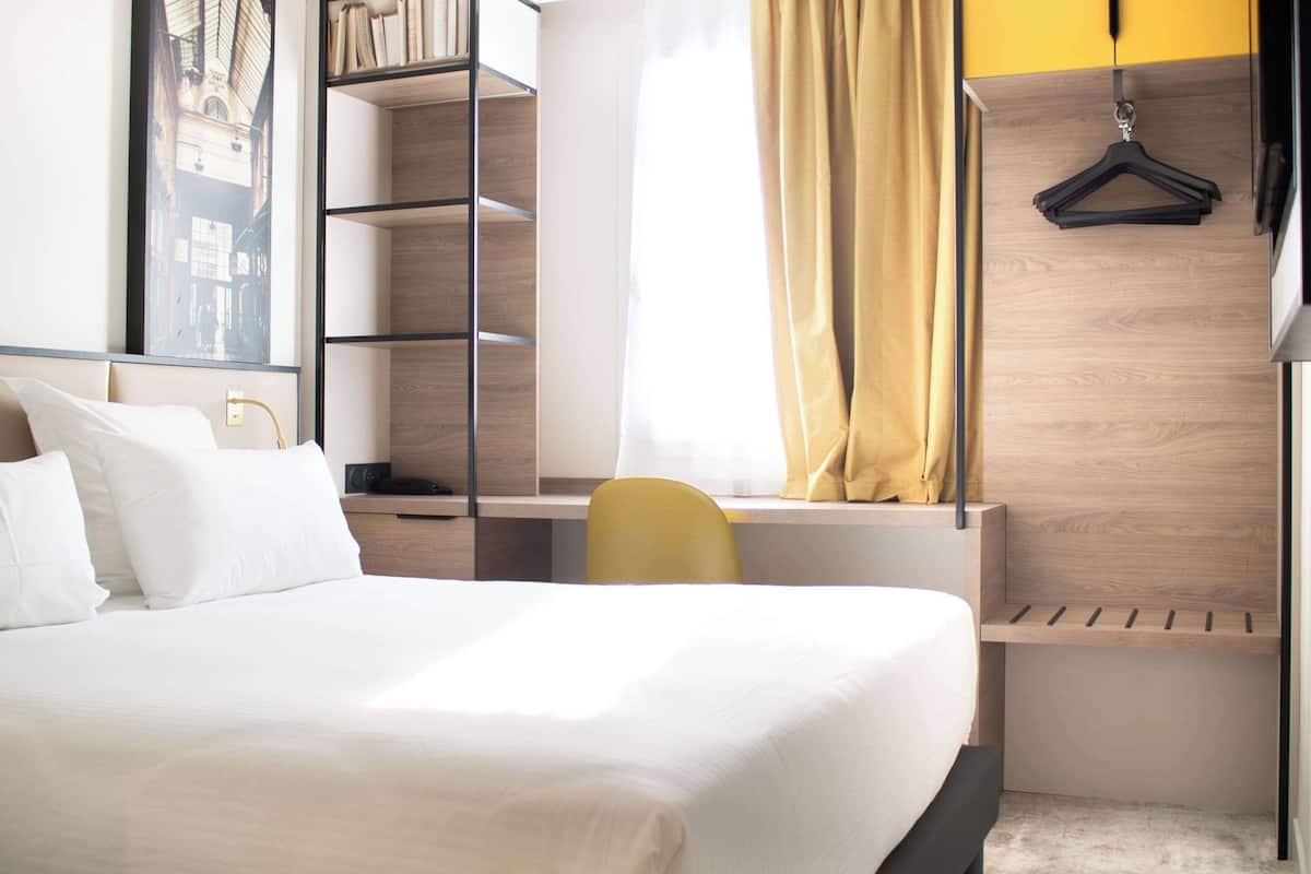 Une chambre d’hôtel moderne équipée d’un lit, d’un bureau, d’une chaise, d’une armoire et d’un porte-manteau.