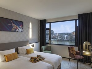 A hotel room with a large bed, a side table, a chair, and a view of the city through a large window. - Mercure Namur Hotel (Namur)