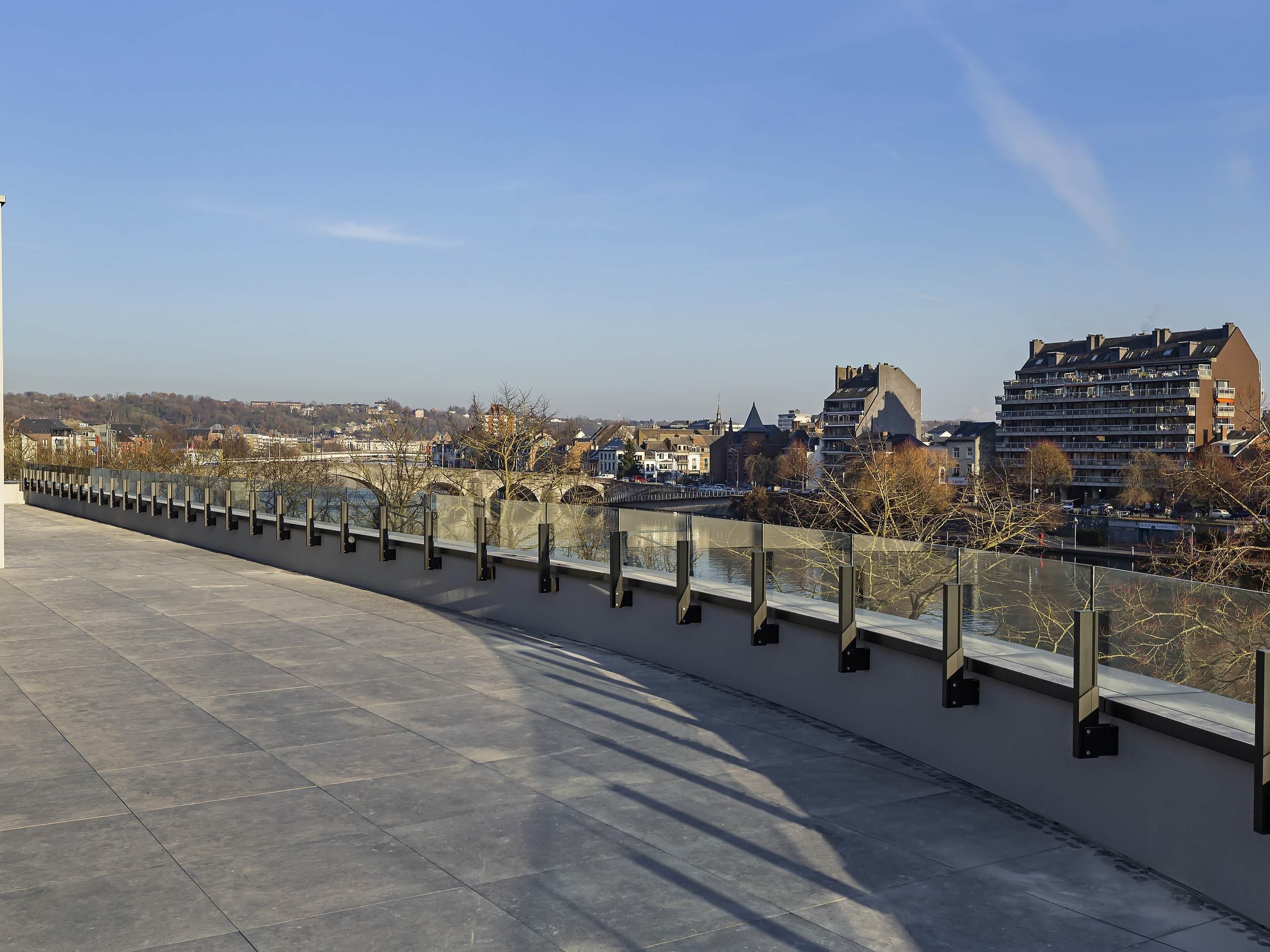 Une promenade au bord de la rivière, agrémentée d’une rambarde moderne, offrant une vue dégagée sur les bâtiments situés de l’autre côté et permettant d’apercevoir un pont au loin.