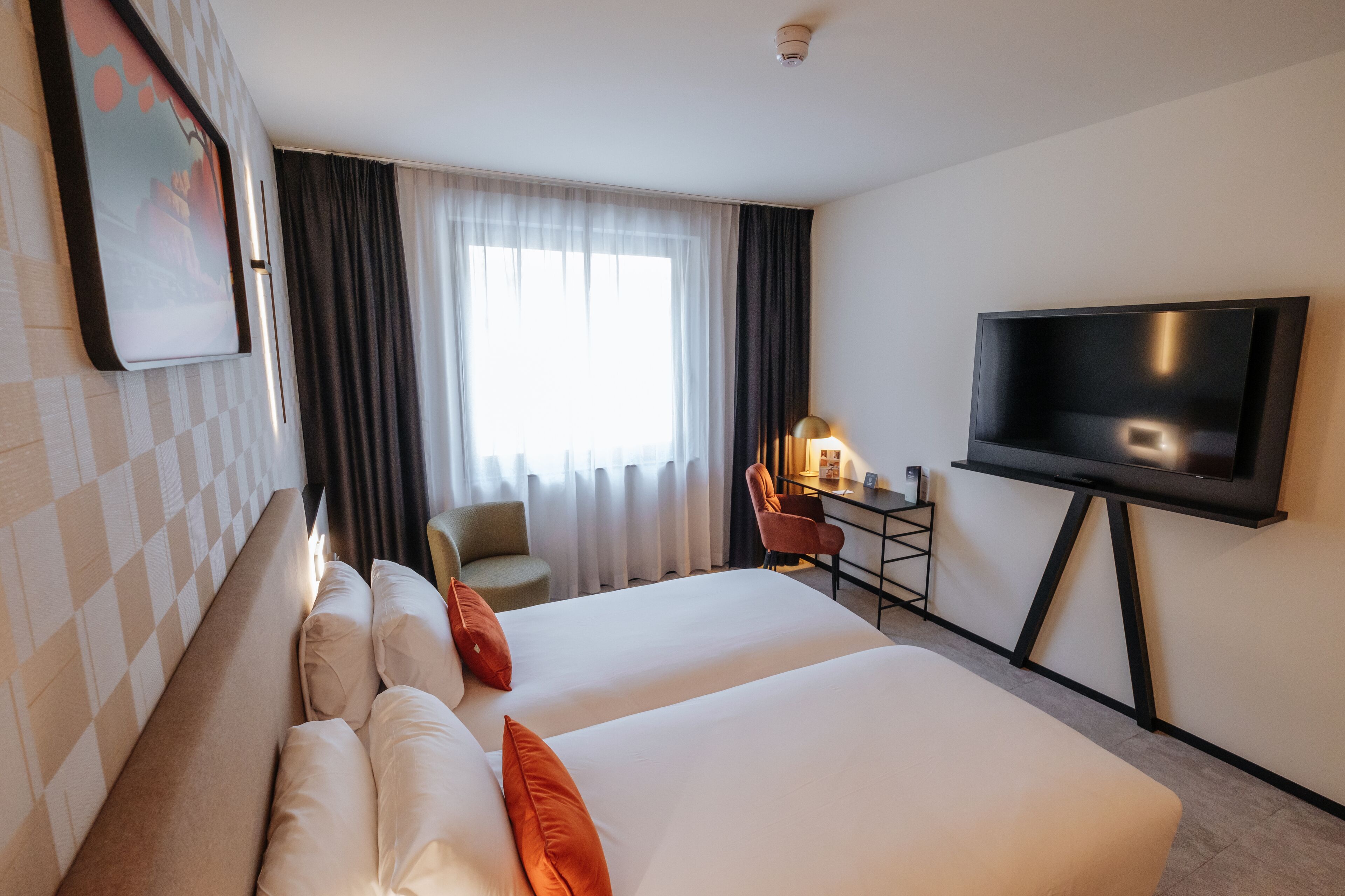 Habitación de hotel con dos camas, un escritorio con silla, un televisor de pantalla plana y un ventanal con cortinas.