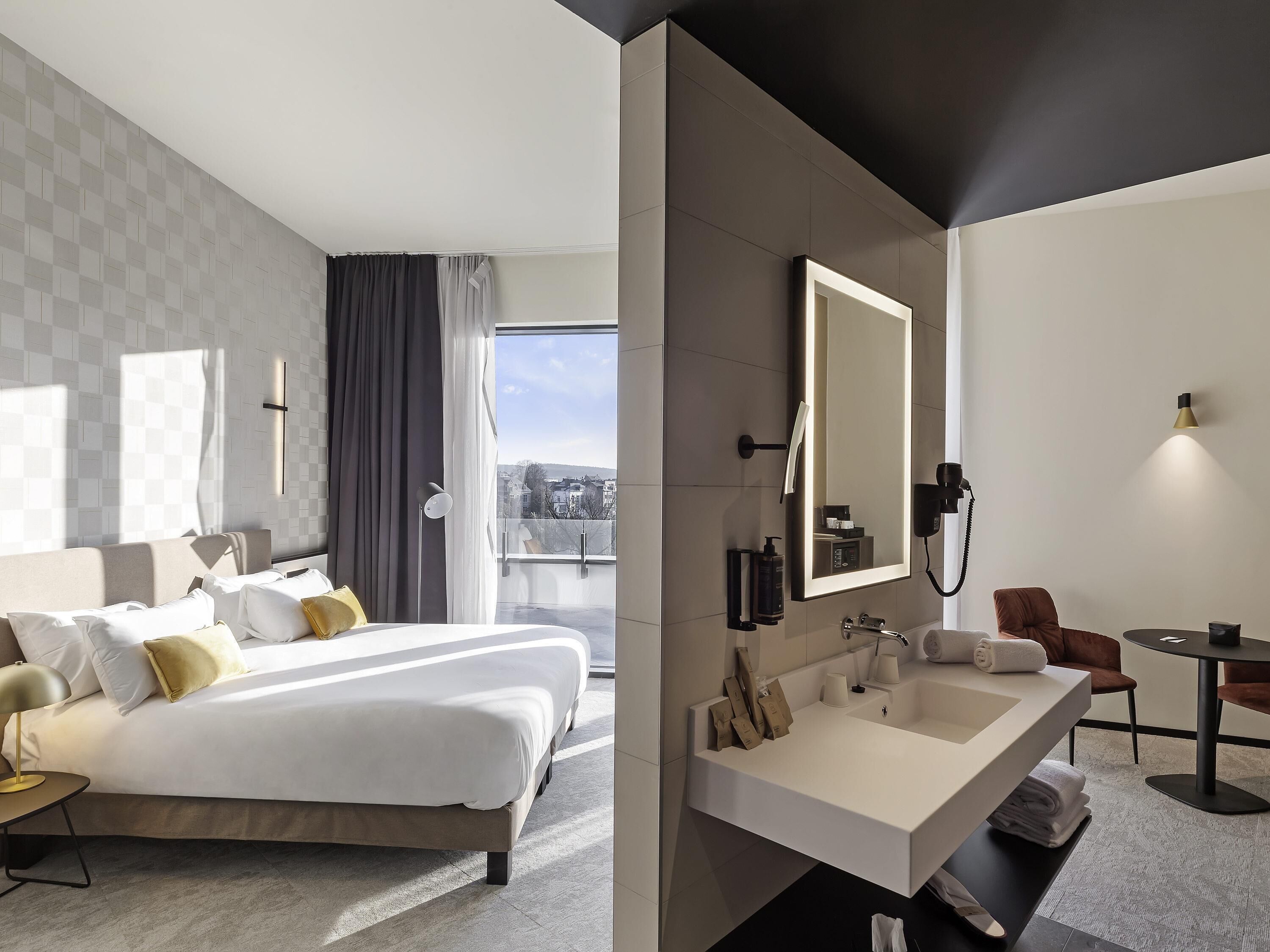 Una habitación de hotel moderna con una cama grande, un baño con un espejo amplio y vistas a la ciudad.