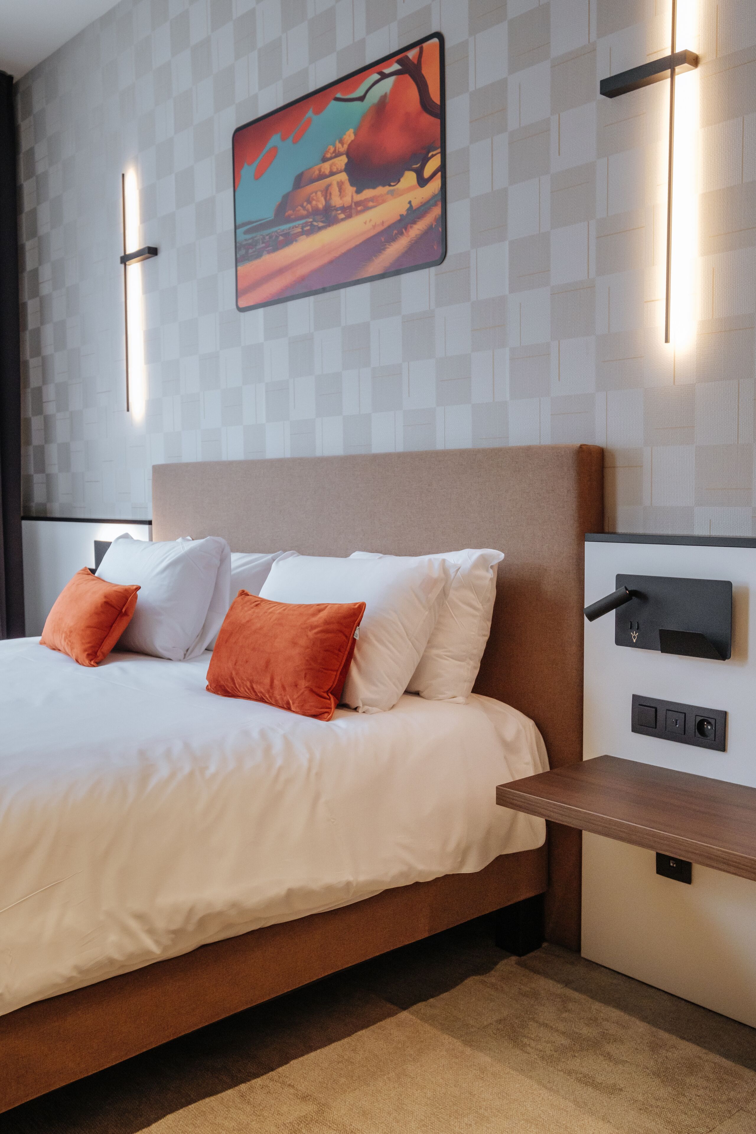 Une chambre d’hôtel moderne avec un grand lit, du linge de lit blanc, des oreillers orange, une tête de lit, une table de chevet, un panneau de commande fixé au mur et un tableau encadré accroché au mur.
