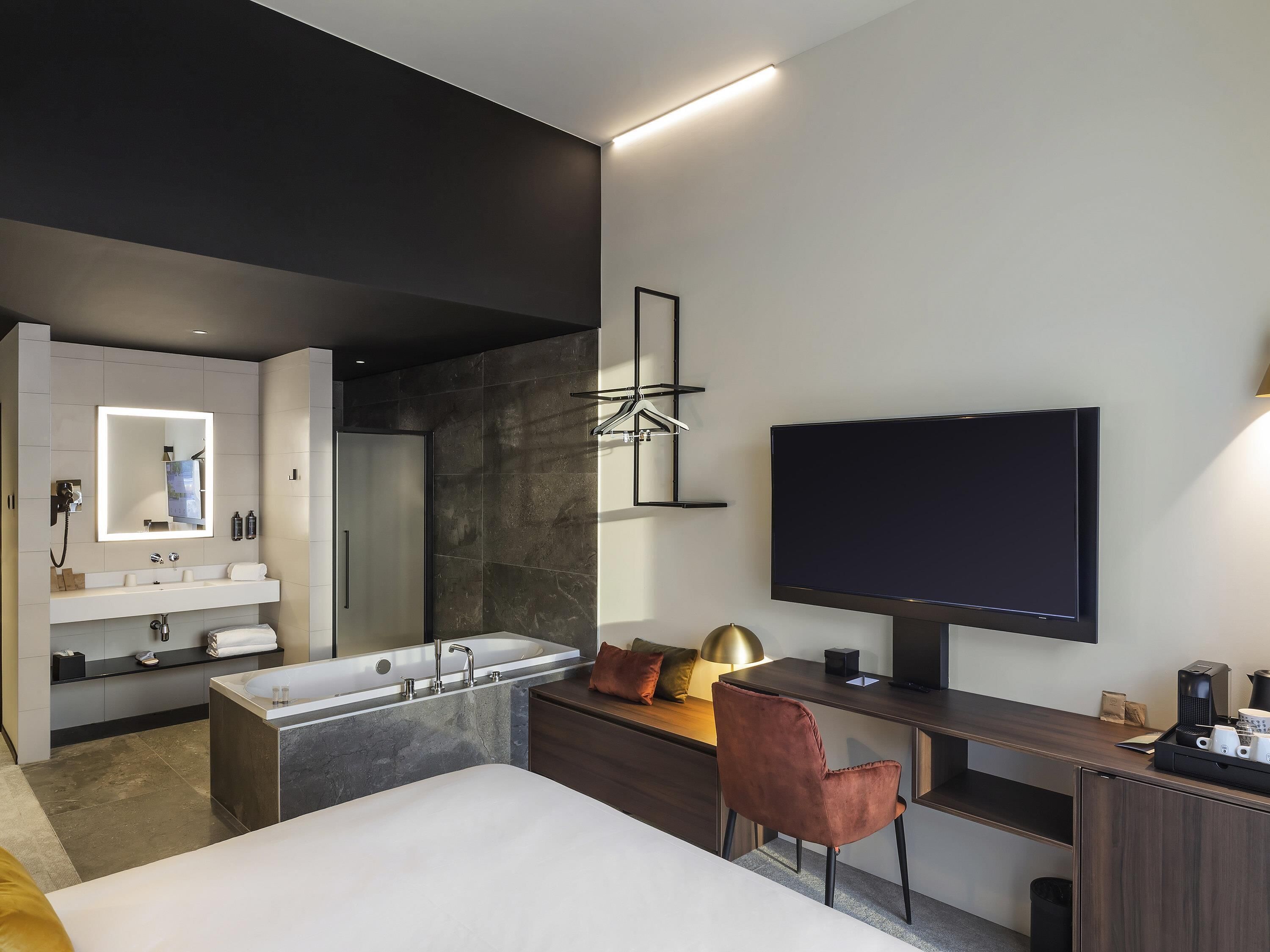 Ein modernes Hotelzimmer mit einem großen Bett, einem Schreibtisch, einem Flachbildfernseher, einem großen Spiegel, einer Badewanne und einer Dusche.