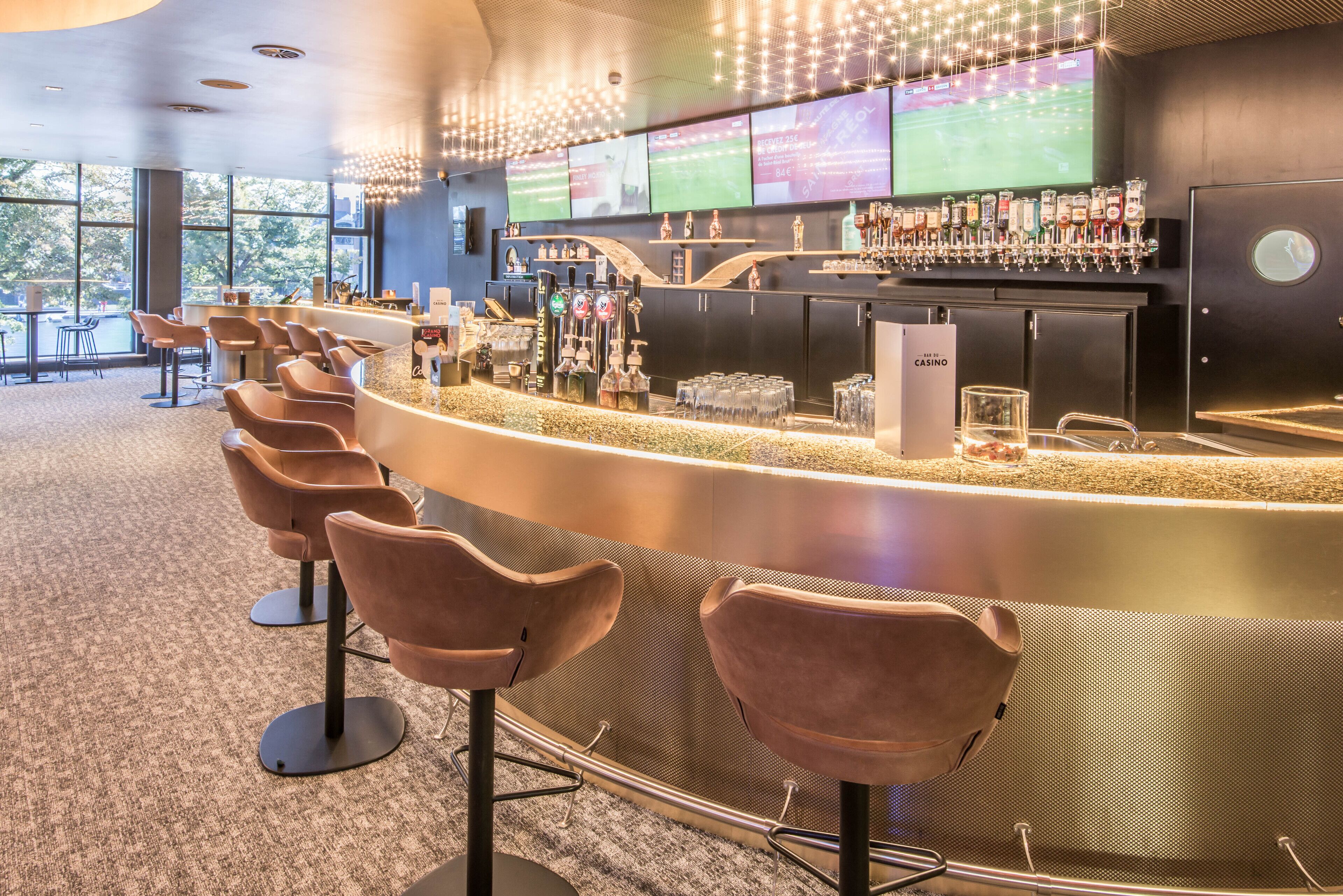 A bar area with a curved counter, multiple taps, and a selection of bottles. There are bar stools and a large screen displaying sports.