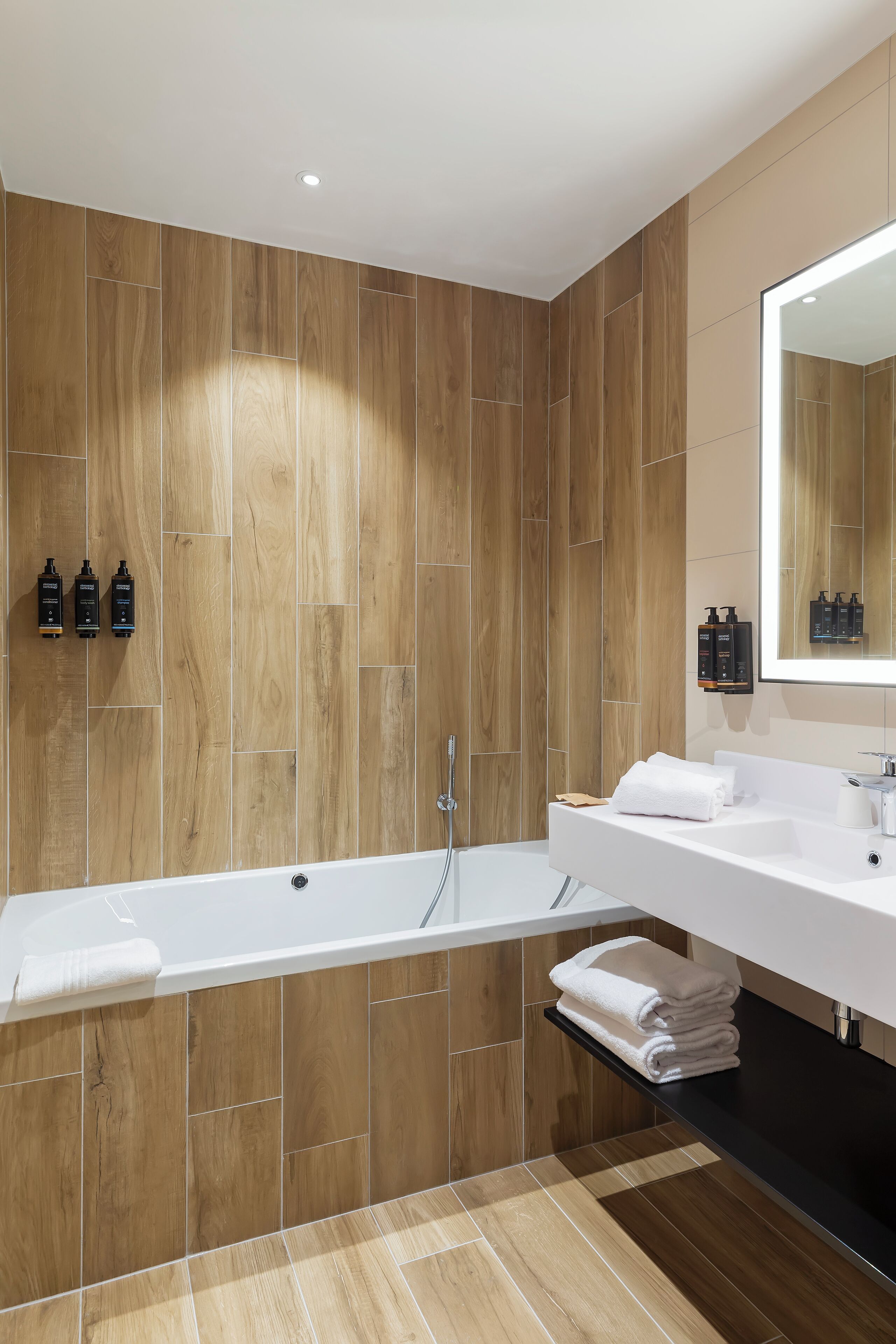 Un baño moderno con pared de madera, una bañera blanca y un espejo grande sobre el lavamanos.